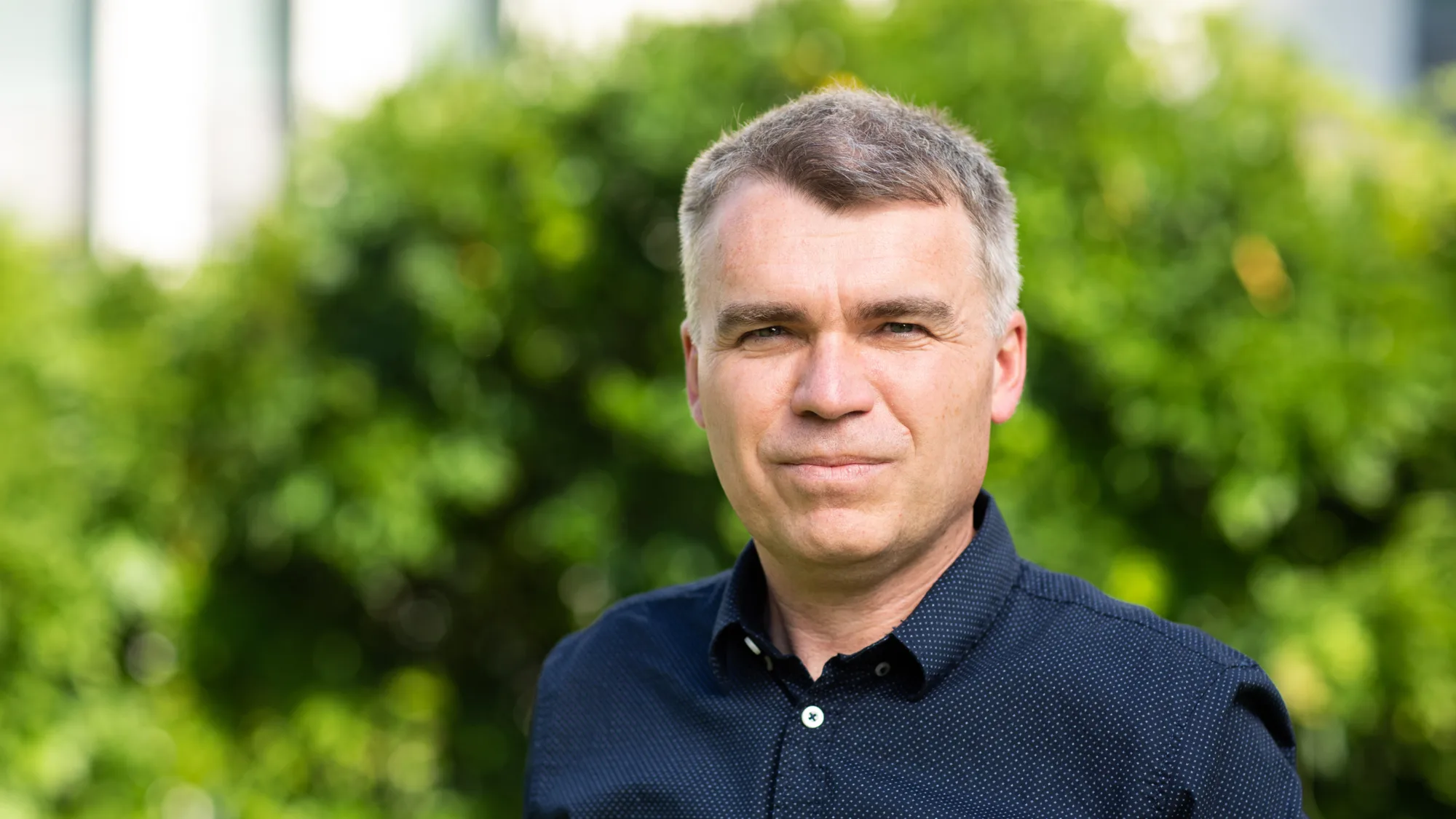 Head and sholders shot of Rasti wearing a dark blue shirt against green foliage