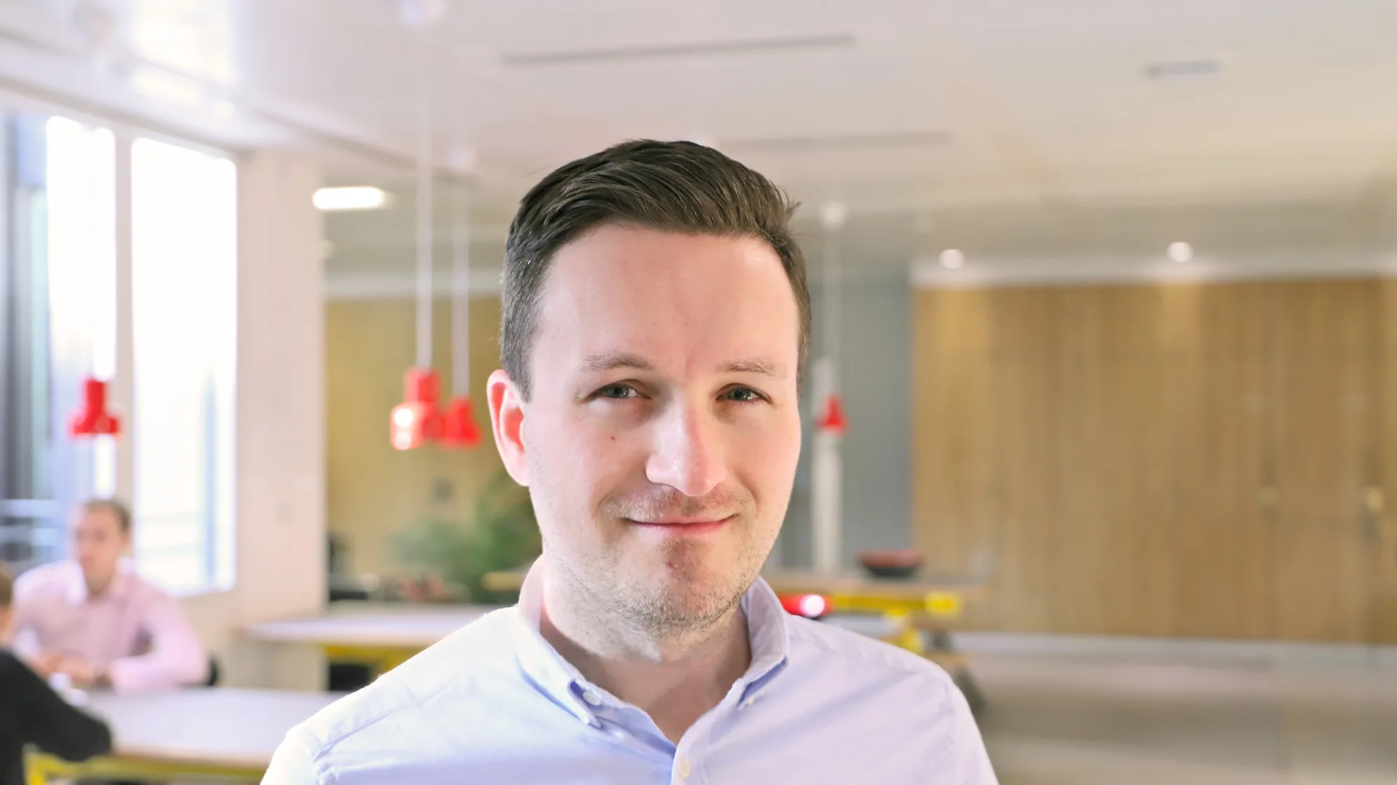 Man with short dark hair wearing a light blue button-up shirt stands in a modern office. Background includes people at tables with blurred faces, wooden elements, red pendant lights, and large windows.