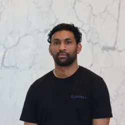 A person with dark curly hair stands against a light marble wall, wearing a black T‑shirt with the word ‘Cundall’ printed in blue on the chest. Upper body is centered in the frame.