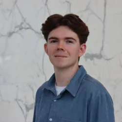 Zac in a blue open collar shirt with a white t-shirt underneath in front of a marbled wall.