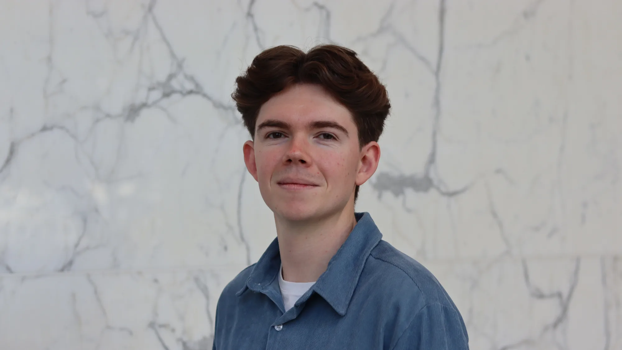 Zac in a blue open collar shirt with a white t-shirt underneath in front of a marbled wall.