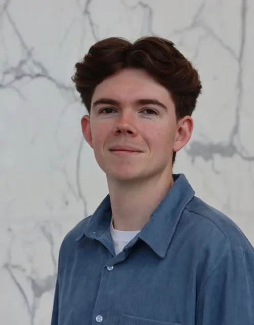 Zac in a blue open collar shirt with a white t-shirt underneath in front of a marbled wall.