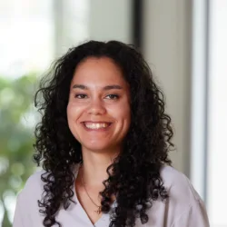 A person with shoulder-length curly dark hair wears a light top and a gold necklace with a round pendant. The softly blurred background includes green foliage and a window, suggesting an indoor setting with natural light.
