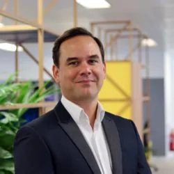 Person wearing a dark suit jacket over a white collared shirt standing indoors. The background shows a modern office space with wooden geometric partitions, green plants, and bright overhead lighting.