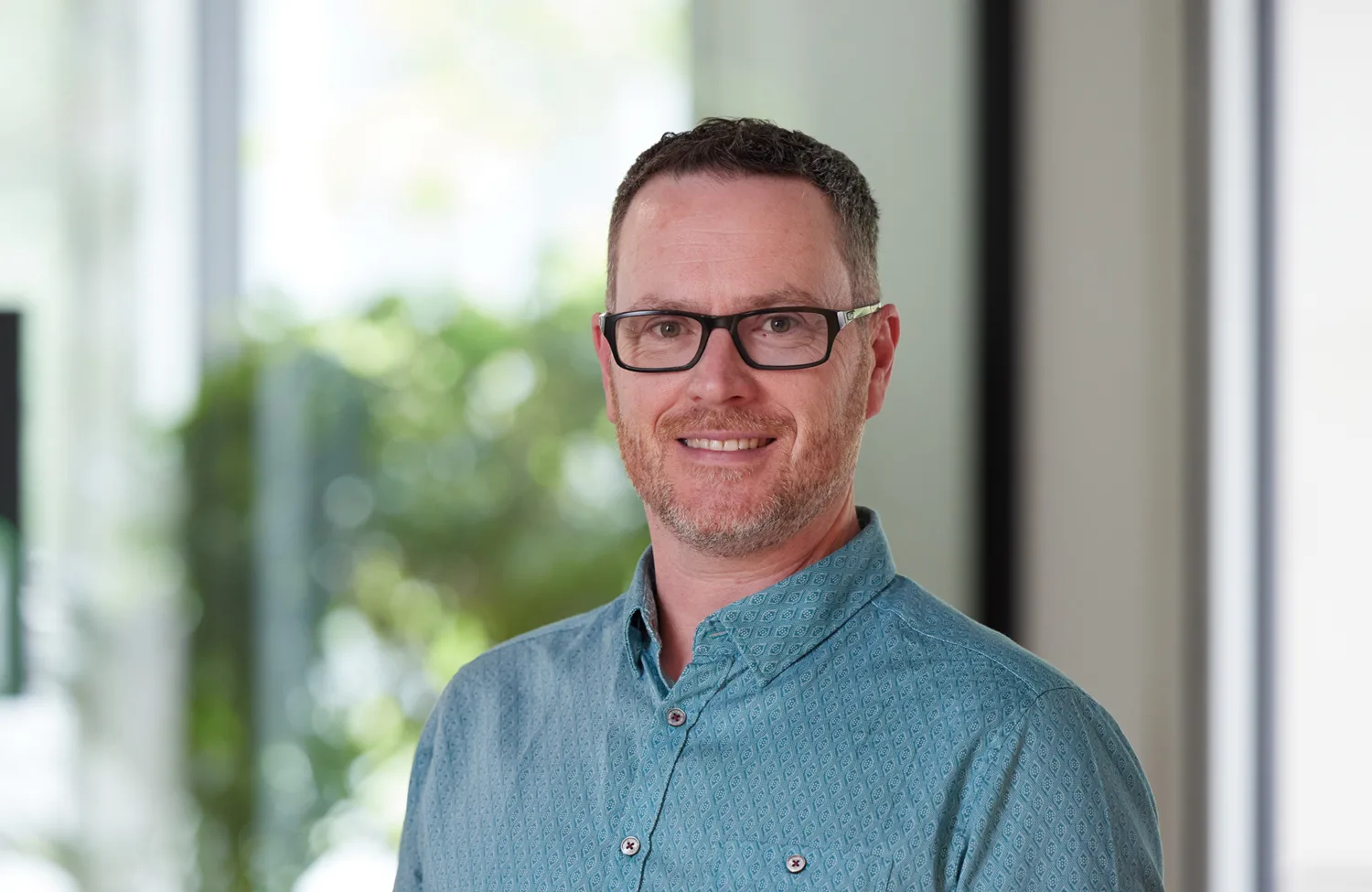 A person with short hair and glasses wearing a blue button-up shirt is seated indoors. Behind them are green plants and large windows with natural light filtering through.