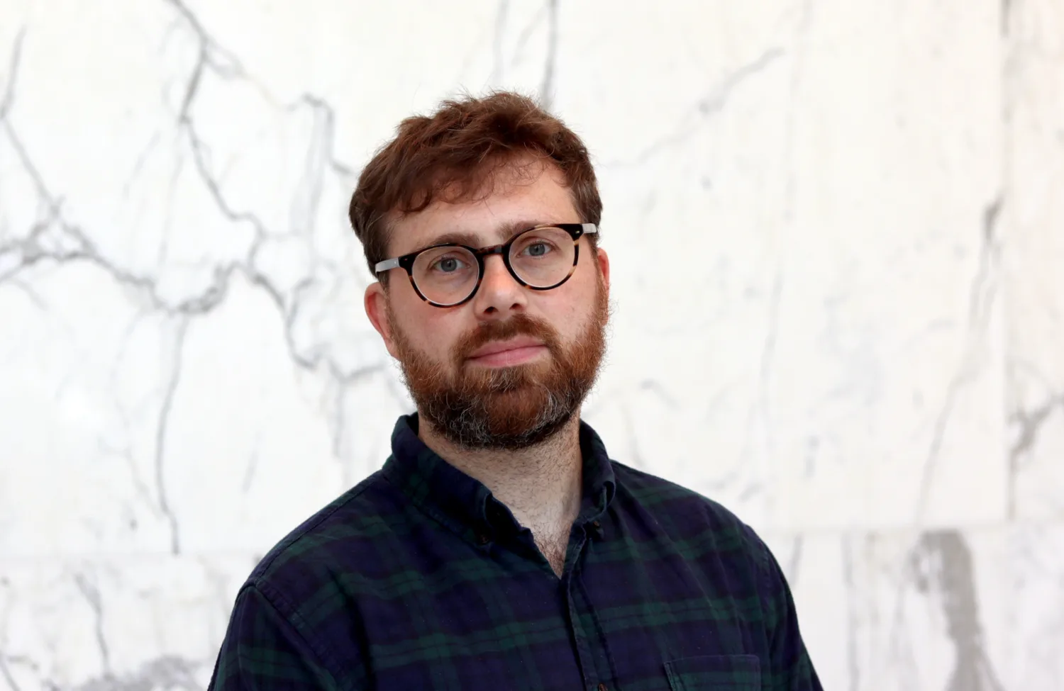 A person with short brown hair and a beard stands in front of a white marble wall with grey veining. They wear a dark green and navy checkered shirt.