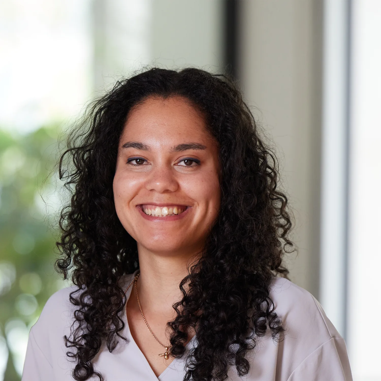 A person with shoulder-length curly dark hair wears a light top and a gold necklace with a round pendant. The softly blurred background includes green foliage and a window, suggesting an indoor setting with natural light.