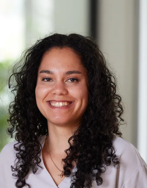 A person with shoulder-length curly dark hair wears a light top and a gold necklace with a round pendant. The softly blurred background includes green foliage and a window, suggesting an indoor setting with natural light.