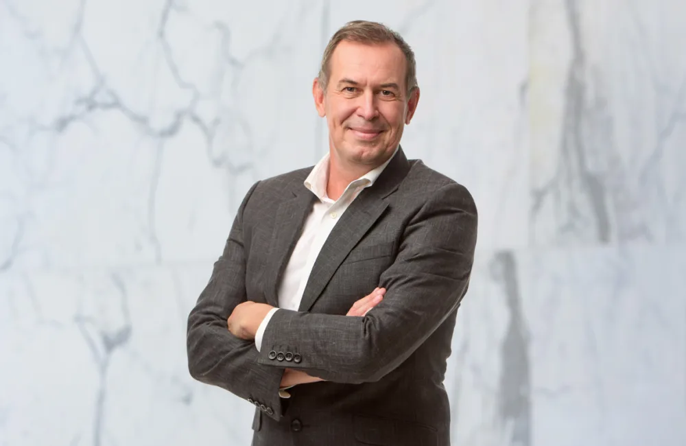A person with short hair wearing a grey suit and white shirt stands with arms crossed in front of a white marble background, facing the camera.