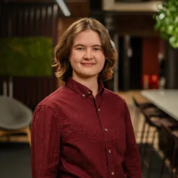 A person with shoulder‑length hair wearing a burgundy button‑up shirt stands indoors near a long table with chairs, lounge seating, brick walls, green wall panel, and plants in the background.