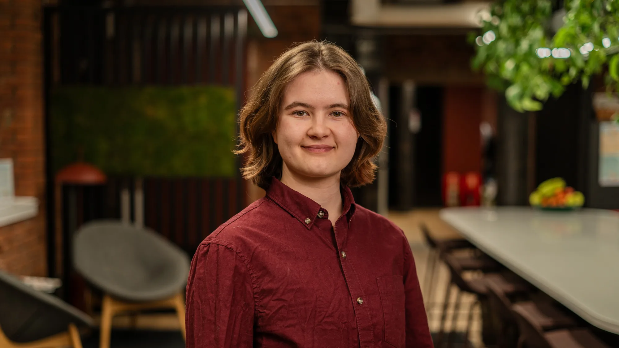 A person with shoulder‑length hair wearing a burgundy button‑up shirt stands indoors near a long table with chairs, lounge seating, brick walls, green wall panel, and plants in the background.
