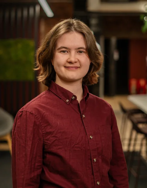 A person with shoulder‑length hair wearing a burgundy button‑up shirt stands indoors near a long table with chairs, lounge seating, brick walls, green wall panel, and plants in the background.
