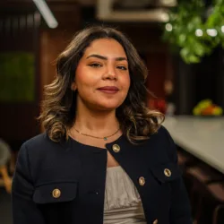 Dana in a dark blazer with gold buttons and a light top stands indoors. Background features a grey chair, green plants, and a long table in a modern lounge area.