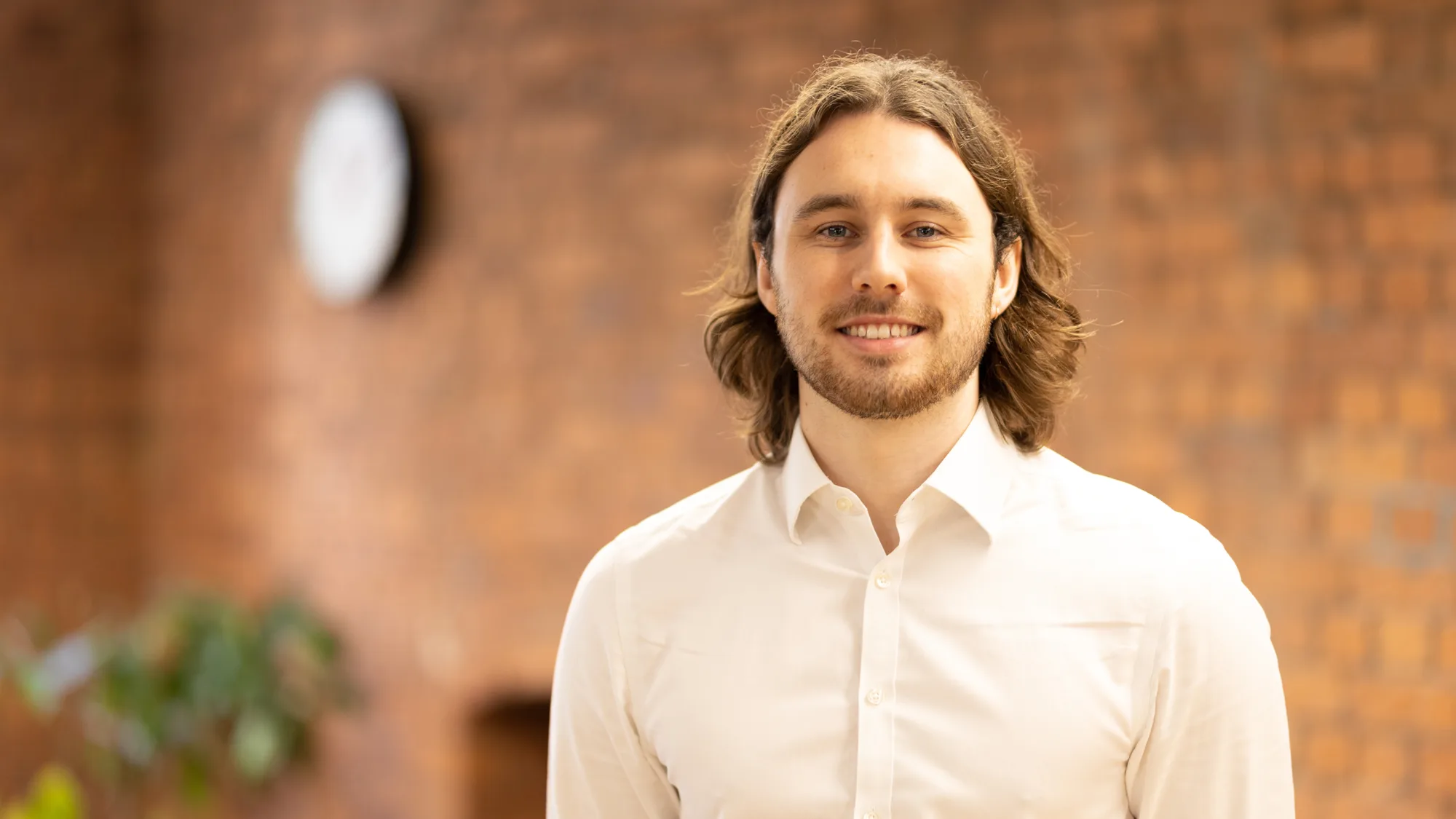 Darnell in an open collar white shirt, smiling to camera in front of a brick wall