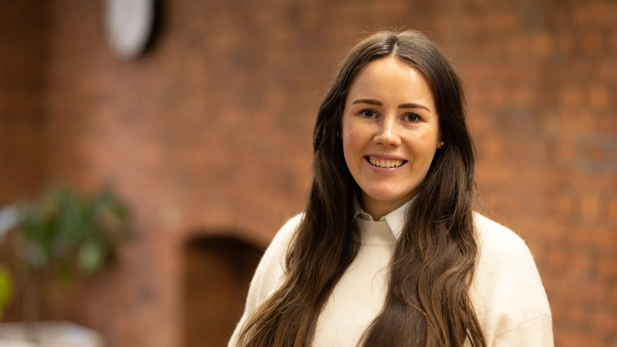 Hannah in a cream jumper and collared shirt, smiling to camera in front of an exposed brick indoor wall