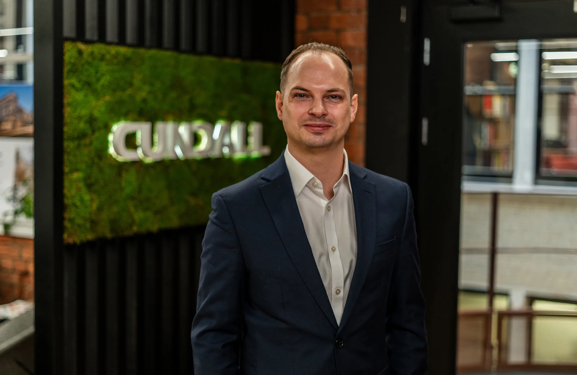 Joe wearing a dark suit jacket and white dress shirt stands indoors near a wall with a green moss panel displaying the text “CUNDALL,” with shelves and a doorway visible in the background.