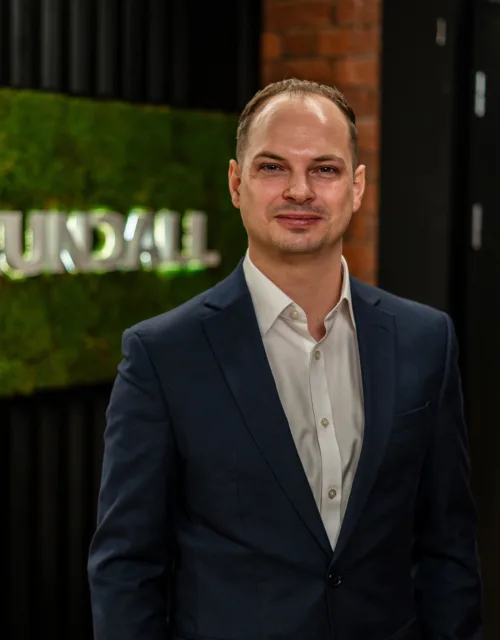 Joe wearing a dark suit jacket and white dress shirt stands indoors near a wall with a green moss panel displaying the text “CUNDALL,” with shelves and a doorway visible in the background.
