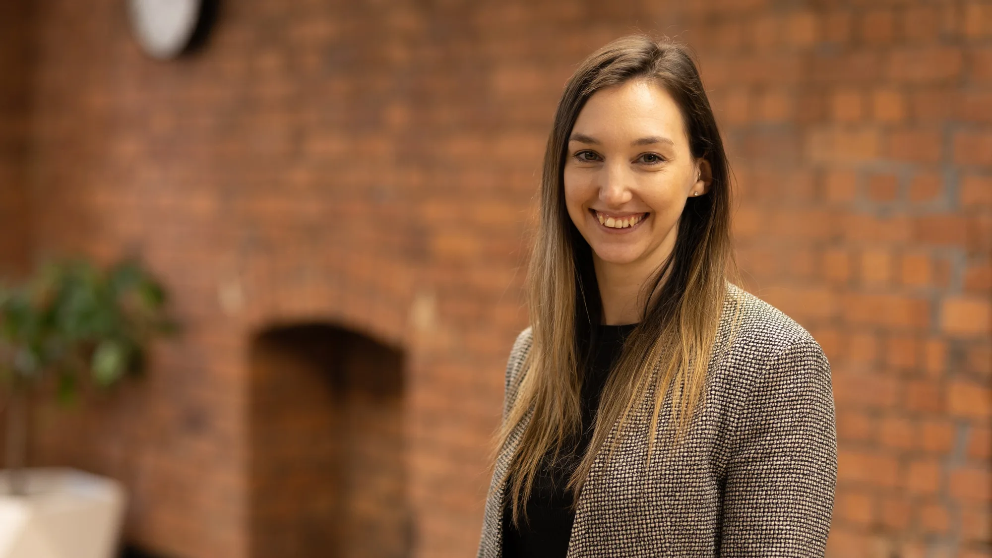 Kimberley stood smiling to camera in front of an exposed brick interior office wall