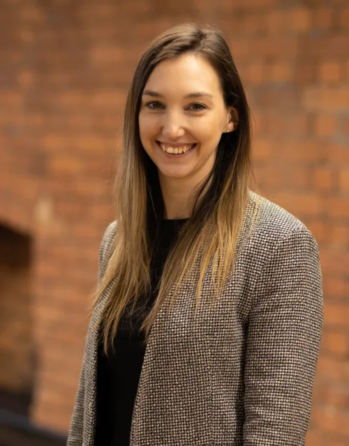Kimberley stood smiling to camera in front of an exposed brick interior office wall