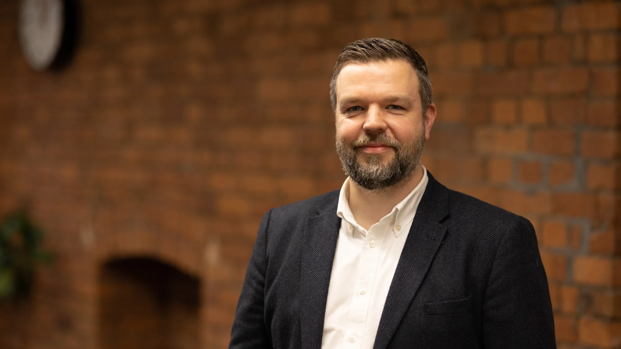 Peter Ridge in a white open collar shirt and suit jacket in front of a brick wall