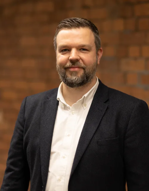 Peter Ridge in a white open collar shirt and suit jacket in front of a brick wall