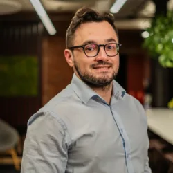 Person wearing a light blue button-up shirt standing indoors. Background includes a long white table with chairs, green plants, a bowl of fruit, and modern ceiling lights in a well-lit office space.