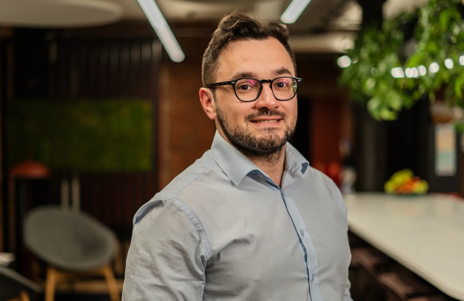 Person wearing a light blue button-up shirt standing indoors. Background includes a long white table with chairs, green plants, a bowl of fruit, and modern ceiling lights in a well-lit office space.