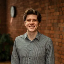 A person wearing a checkered long-sleeve shirt stands indoors in front of a brick wall, with a clock and a leafy plant visible in the background.