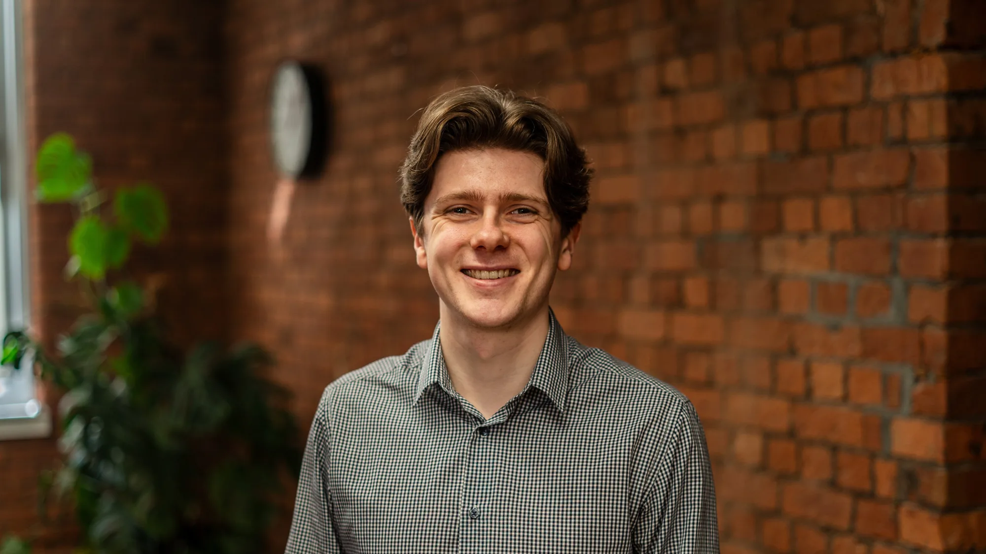 A person wearing a checkered long-sleeve shirt stands indoors in front of a brick wall, with a clock and a leafy plant visible in the background.