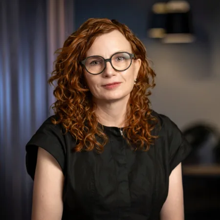 A person with shoulder-length curly red hair wearing a short-sleeved black top is seated indoors. The background shows dark vertical panels, two pendant lights, and blurred plants.