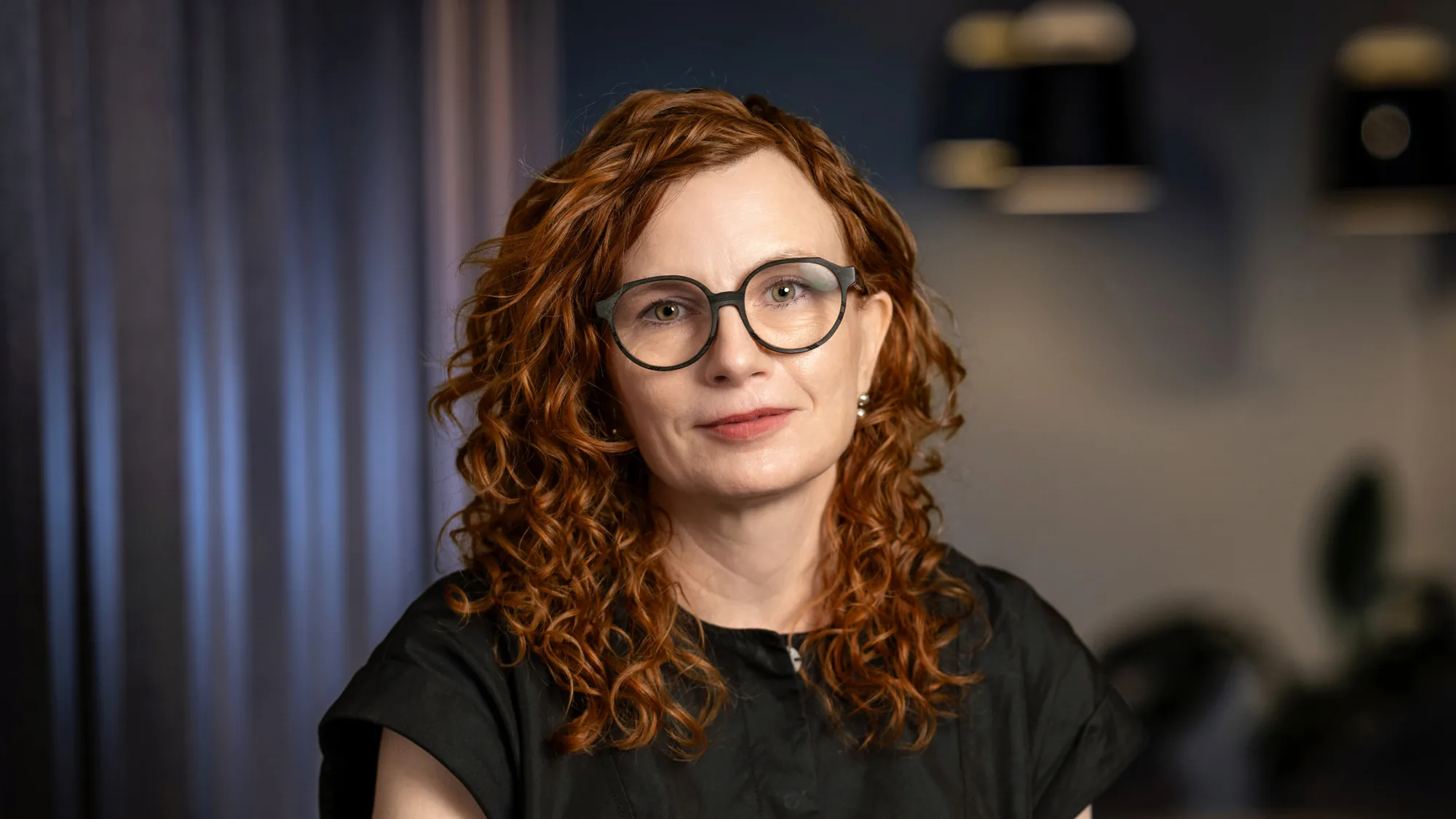 A person with shoulder-length curly red hair wearing a short-sleeved black top is seated indoors. The background shows dark vertical panels, two pendant lights, and blurred plants.