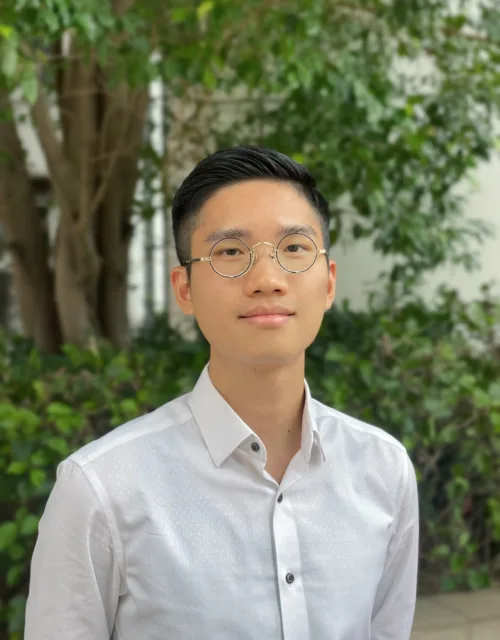 A person wearing round metal-framed glasses and a white button-up shirt is photographed from the chest up, standing outdoors in front of blurred green foliage and a tree trunk in soft natural light.