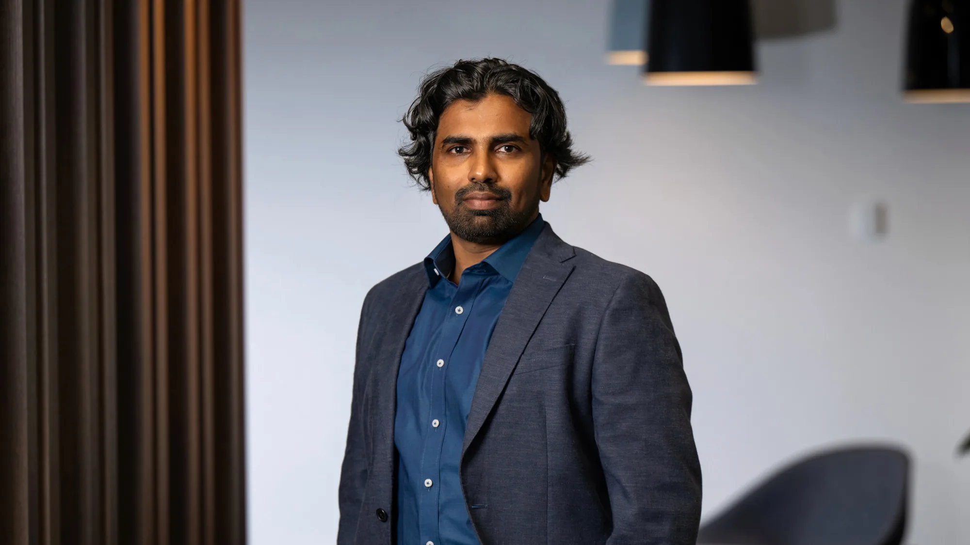 Nanda wearing a dark blue shirt with a dark grey suit jacket standing with arms in pocket in an office setting