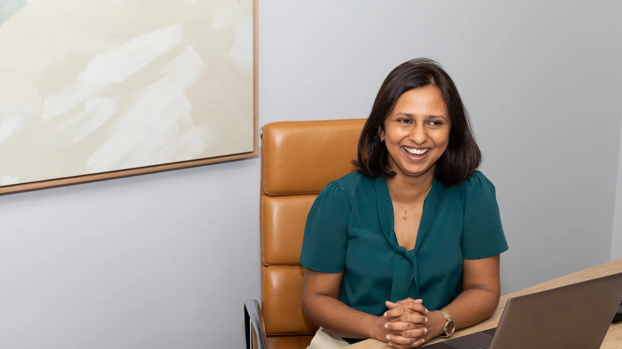 A person sits at a desk in a modern office, wearing a short‑sleeved teal blouse and clasping their hands. A laptop is open on the wooden desk, and a large abstract painting hangs on the wall behind them.