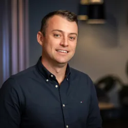 A person wearing a dark long-sleeved button-up shirt with a small embroidered logo on the chest. The background includes vertical panels, two pendant lights, and blurred plants.