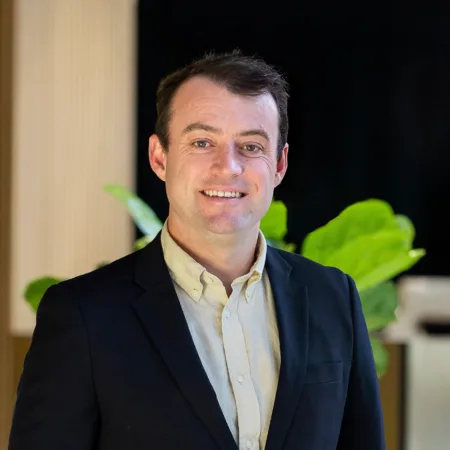 Nathan in a dark blazer and light button-up shirt stands before vertical wooden panels and green plants.