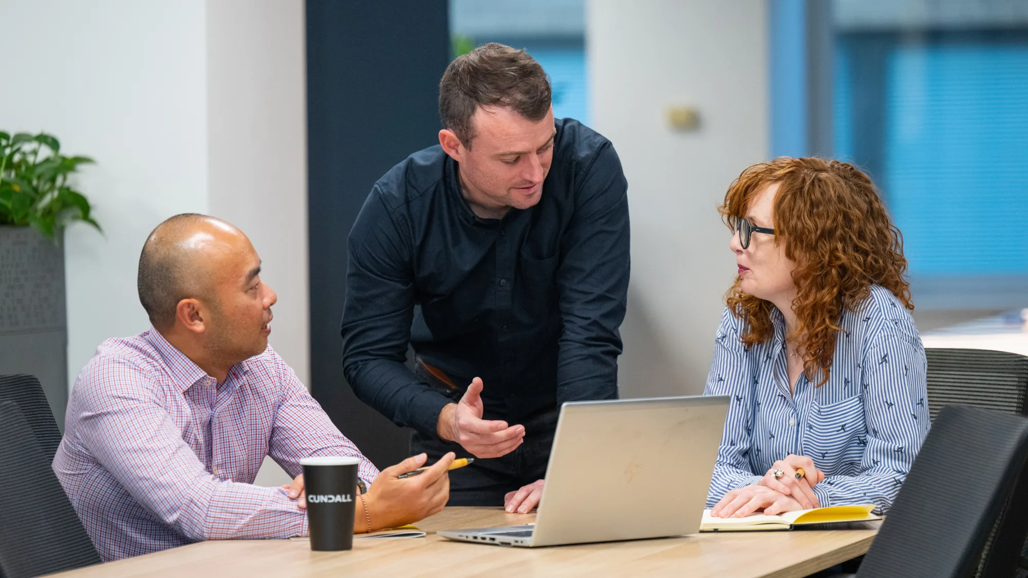 Three individuals in an office setting are engaged in discussion around a table; one stands and gestures, while the others sit—one with a laptop and notebook, the other holding a "CUNDALL"-branded coffee cup.