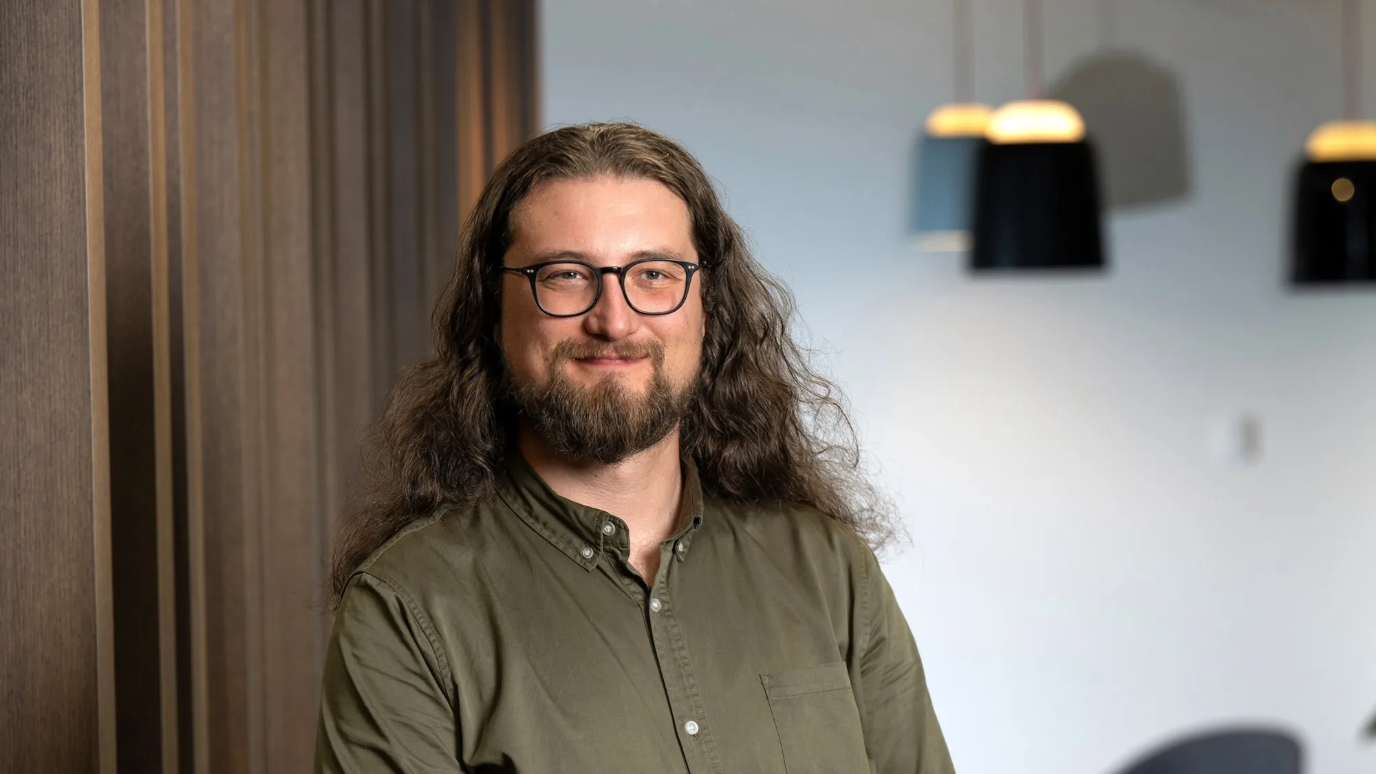 Simon wearing a khaki shirt and smiling to camera stood in front of wooden panelling in the office
