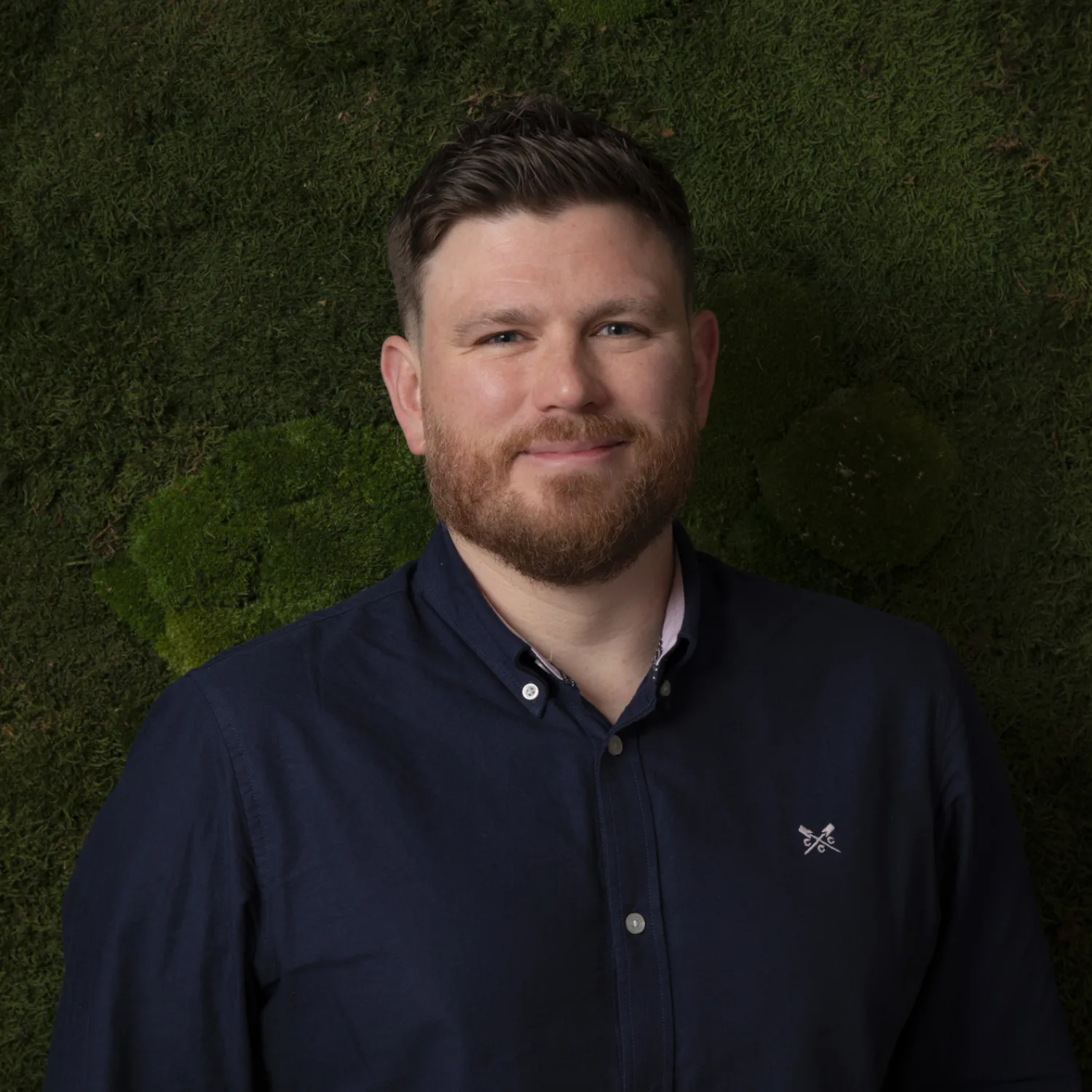 A person wearing a dark navy button-up shirt with a small embroidered logo on the left chest stands against a textured green moss wall background. The shirt is fully buttoned except for the top button.