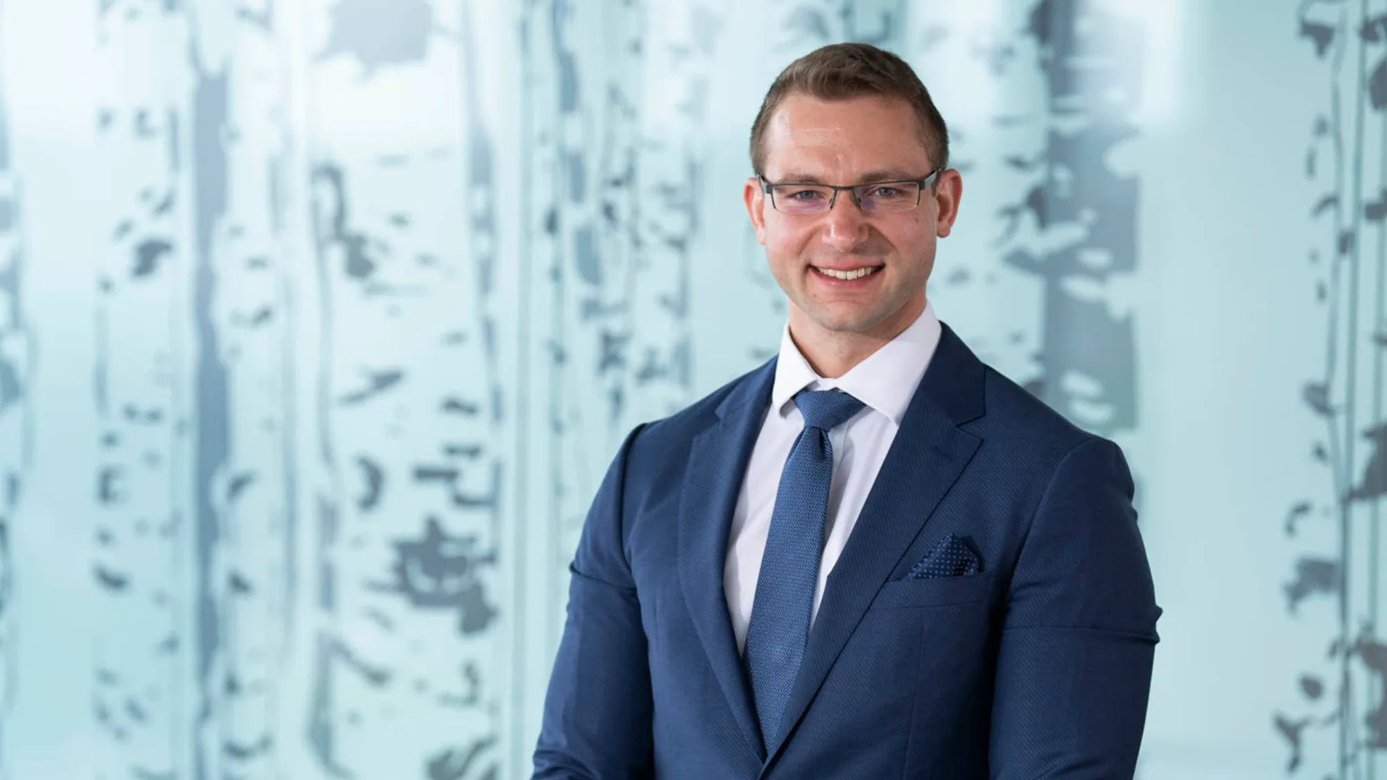 Athanasios wearing a dark suit, white shirt and blue tie, smiling to camera in front of a frosted glass screen.