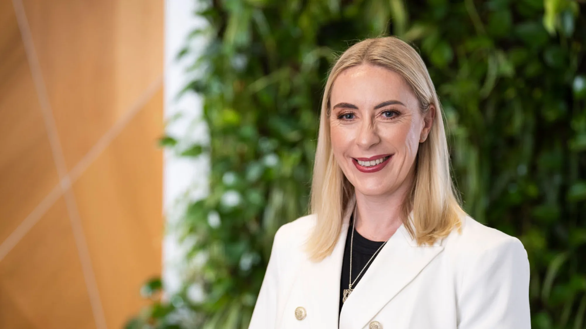Carole smiling in white double breasted jacket in front of a green living wall