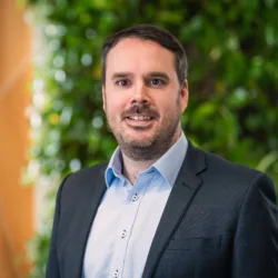 Colin looking to camera in a pale open collar shirt and suit jacket in front of a green plant wall