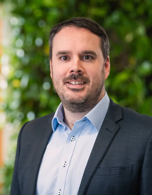 Colin looking to camera in a pale open collar shirt and suit jacket in front of a green plant wall