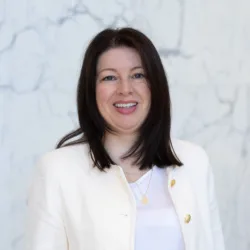 A person wearing a white textured jacket with gold buttons over a white top stands in front of a light grey marble wall. The jacket is open, and two layered necklaces with small pendants are visible.