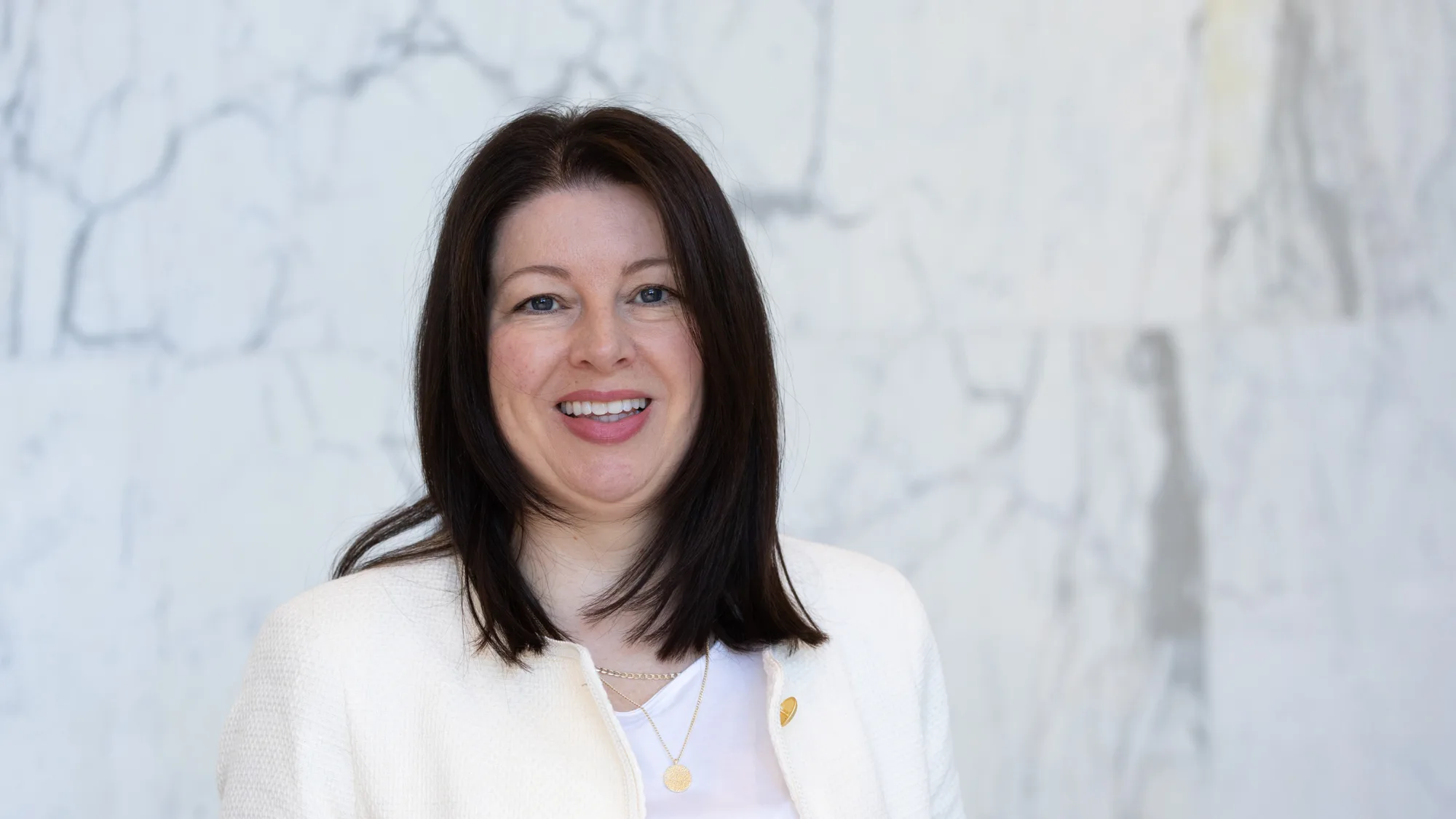 A person wearing a white textured jacket with gold buttons over a white top stands in front of a light grey marble wall. The jacket is open, and two layered necklaces with small pendants are visible.