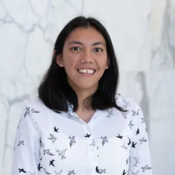 Hannah standing against a light gray marble wall, wearing a white button-up shirt patterned with black bird silhouettes. She has shoulder-length dark hair and is centered in the frame.