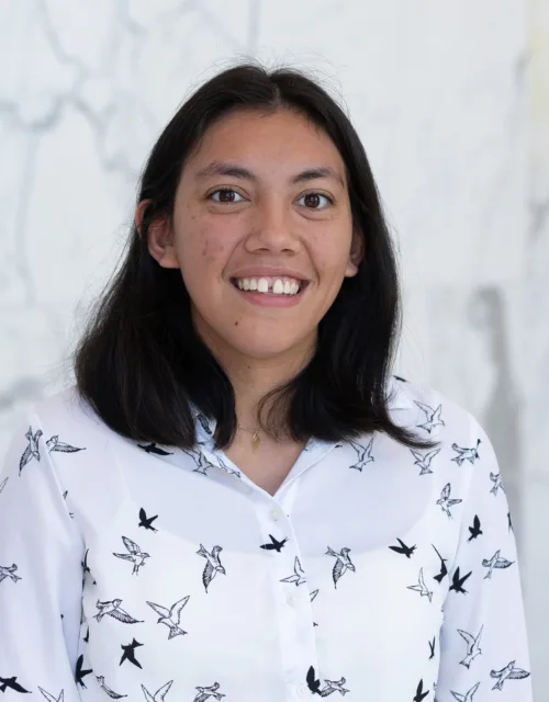 Hannah standing against a light gray marble wall, wearing a white button-up shirt patterned with black bird silhouettes. She has shoulder-length dark hair and is centered in the frame.