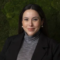 A person wearing a dark blazer over a grey turtleneck top is positioned against a textured green wall with moss-like plants. The outfit includes pearl earrings visible on both sides.