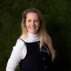 A person with long blonde hair stands against a textured green moss wall, wearing a white long‑sleeve top layered under a dark button‑front pinafore dress, with one arm positioned behind their back.
