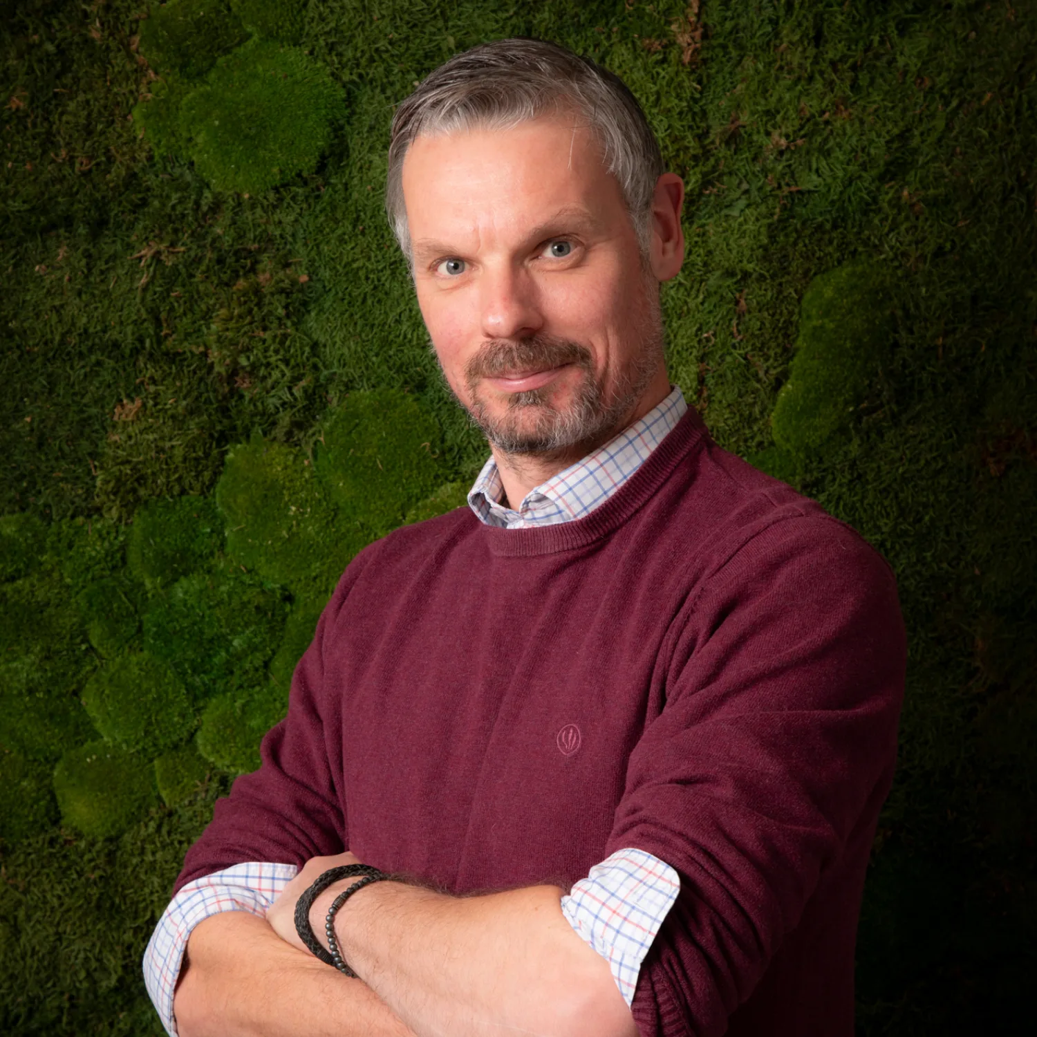 A person wearing a burgundy sweater over a checked shirt stands with folded arms in front of a textured green moss wall.